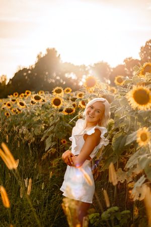 Smiling blonde woman in white dress standing in a sunflower field during golden hour sunset.