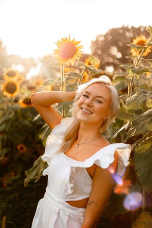 Smiling blonde woman in white ruffled top posing in a sunflower field during golden hour sunset.