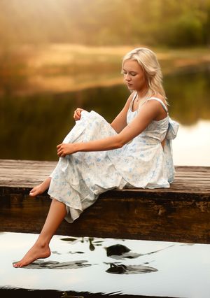 Blonde woman in floral dress sitting barefoot on wooden dock by calm lake in golden sunset light.