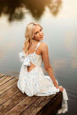 Blonde woman in floral sundress with bow sits on wooden dock by calm reflective lake at golden hour.