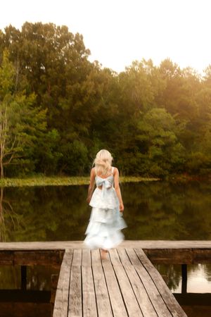 Young girl in tiered light blue dress with bow stands on wooden dock overlooking calm reflective lake surrounded by trees.