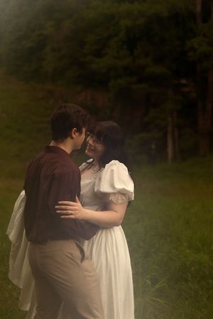 Couple in period costumes embrace outdoors, woman in white dress and man in dark jacket, standing on green grass.