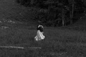 A child in a white dress walks through a grassy field near trees in a black and white photo.