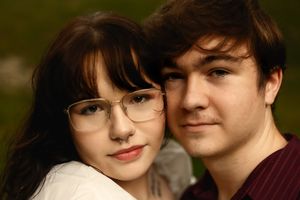 A young couple poses closely together outdoors, the woman wearing glasses and the man in a dark red shirt.