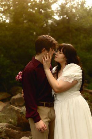 Couple in romantic outdoor setting, woman in white dress touching man's face, surrounded by lush greenery at golden hour.