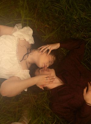 Couple lying in green grass, sharing a tender moment, woman in white dress and man in burgundy shirt.