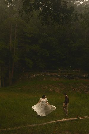 Couple in white dress and hat walking on a log in a lush green forest clearing at dusk.