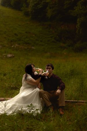 Couple in vintage attire sit on grass toasting with champagne glasses in a lush green hillside setting.