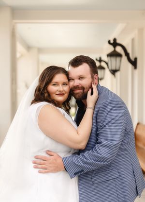 A bride in a white dress and veil embraces her groom in a blue plaid suit, smiling together in an elegant hallway.