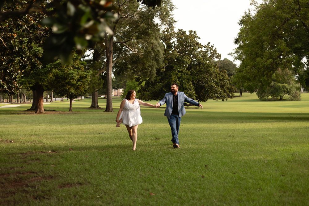 Couple holding hands running through lush green park on wedding day, bride in white dress, groom in blue suit.
