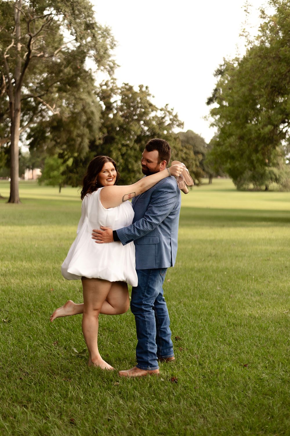 A couple embraces and laughs together on a lush green lawn, woman in white dress, man in blue suit and jeans.