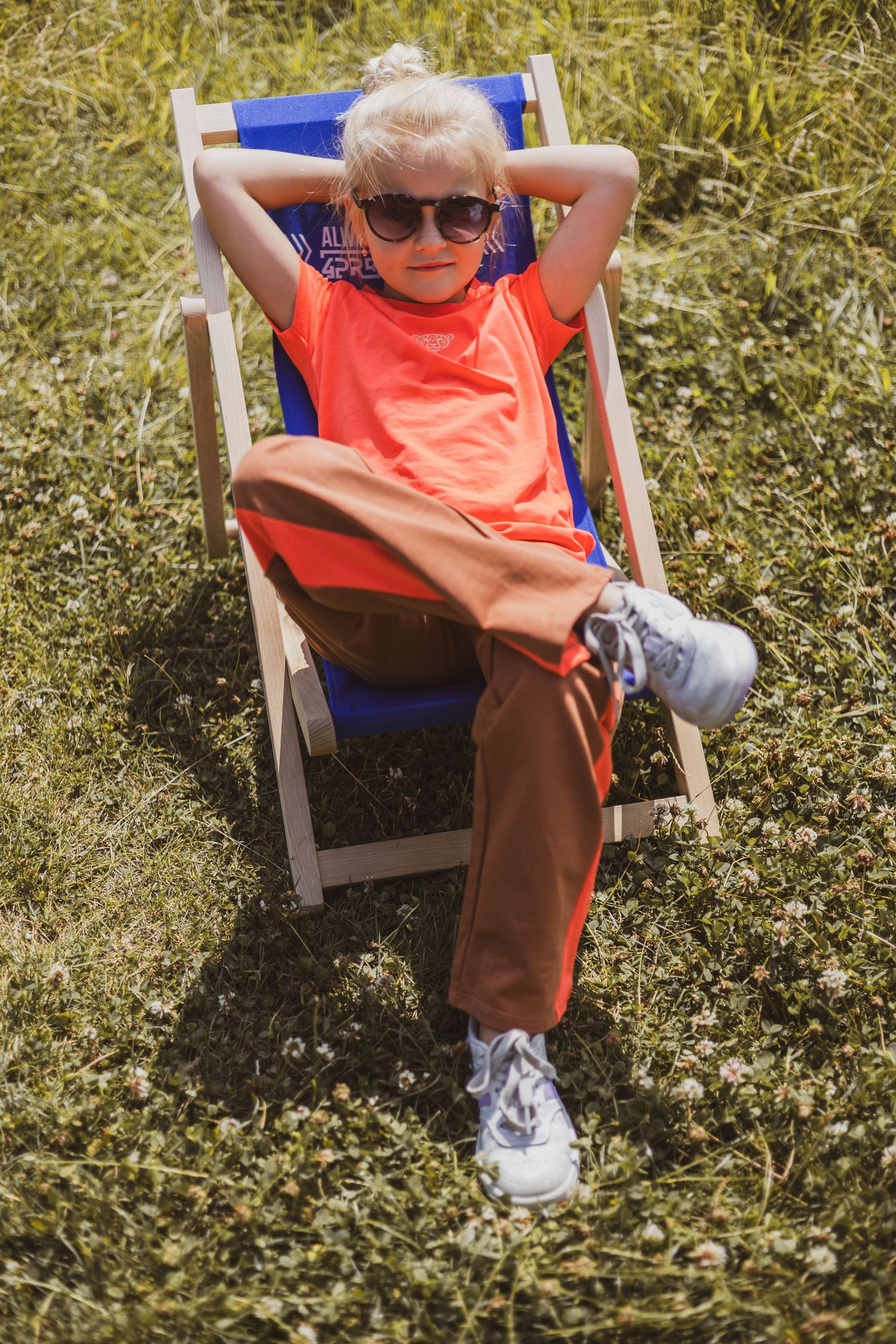Young blonde child relaxing in a blue deck chair on grass, wearing sunglasses and orange shirt on a sunny day.