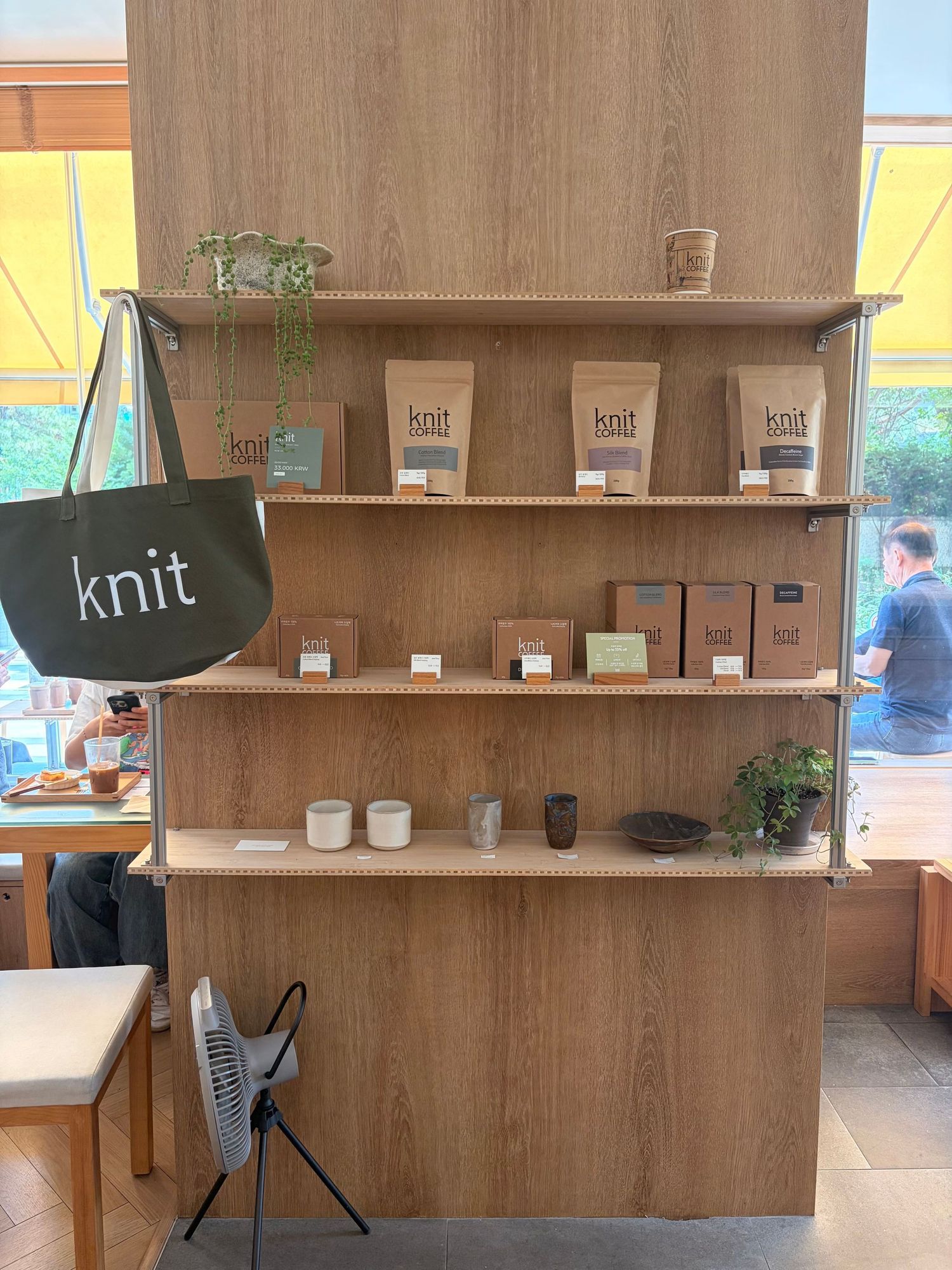 Knit coffee shop retail display shelf with branded bags, coffee packages, and ceramic mugs on wooden shelving unit.