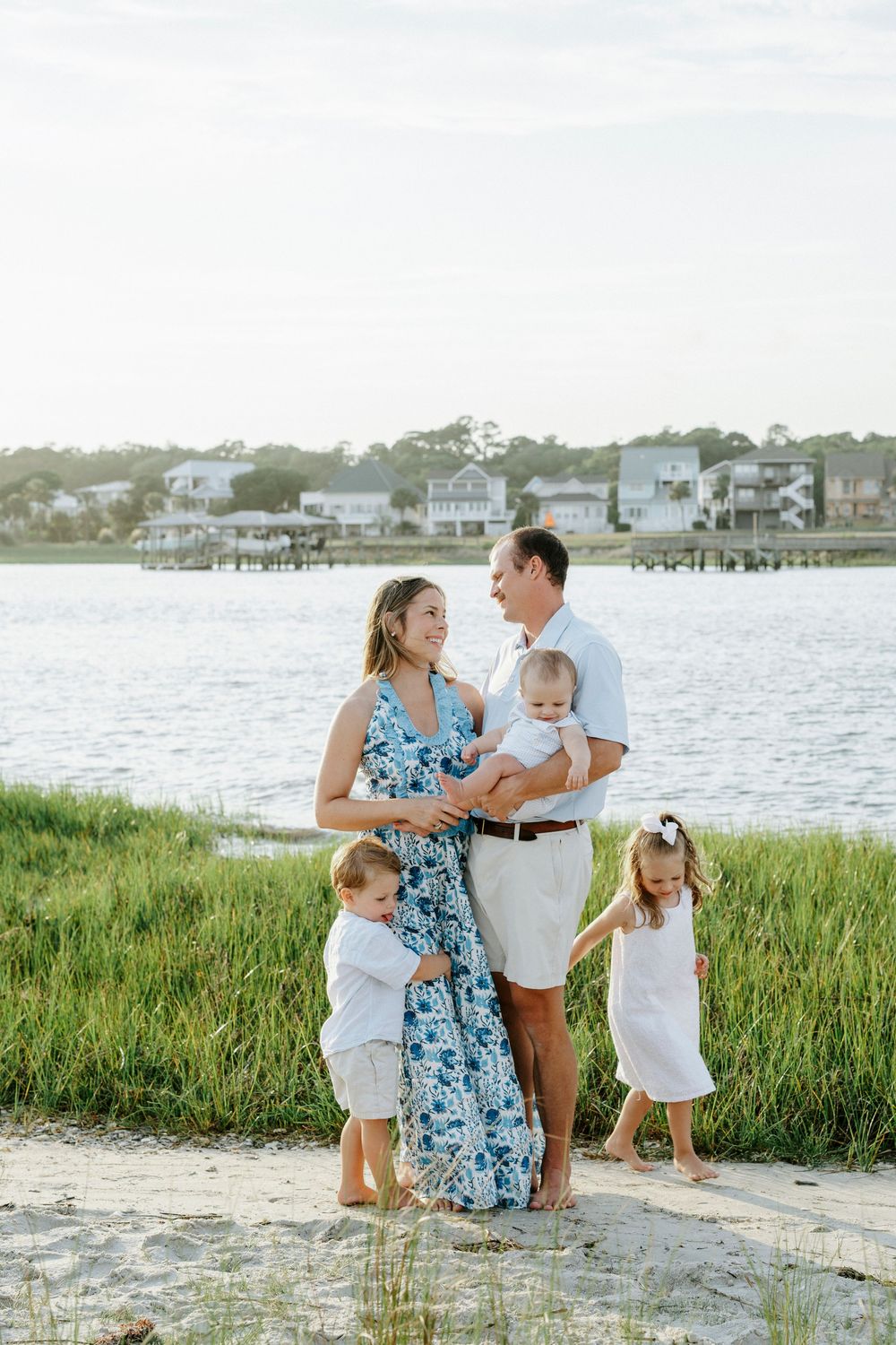 Family of five posing on sandy beach by calm water, parents smiling at each other while holding baby and two young children.