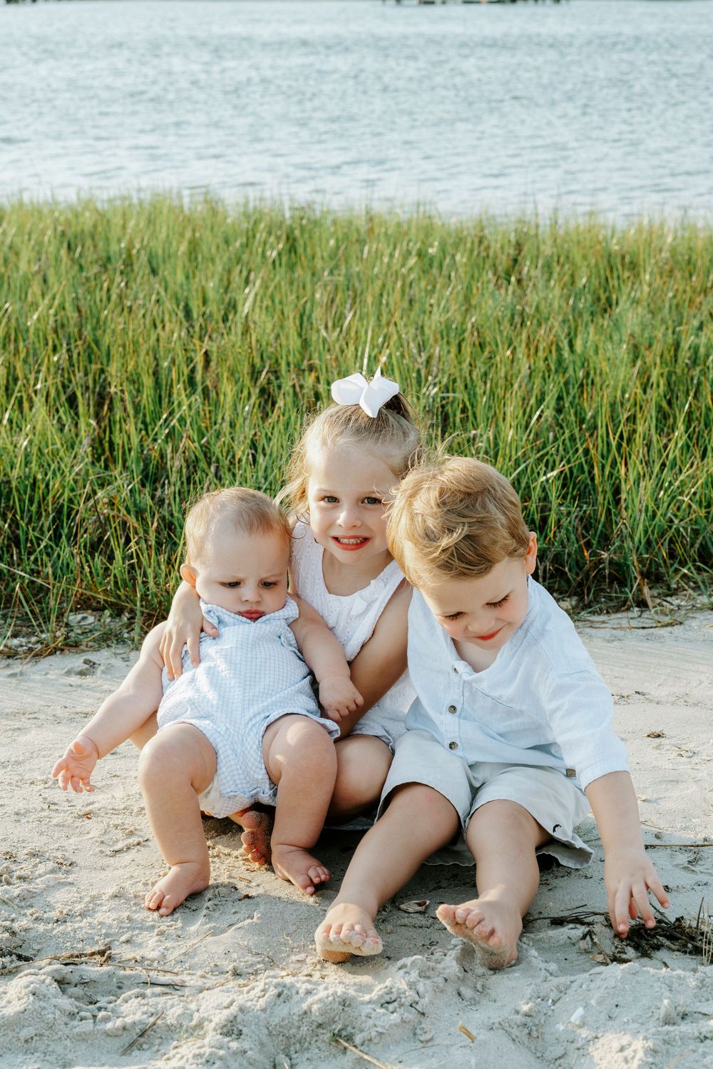 Three young siblings sit together on sandy beach with marsh grass and water backdrop.