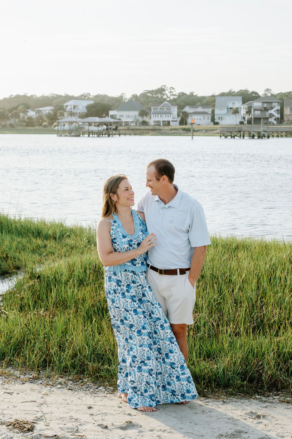 Couple in blue floral dress and polo shirt embracing by a coastal waterway with marsh grass at golden hour.