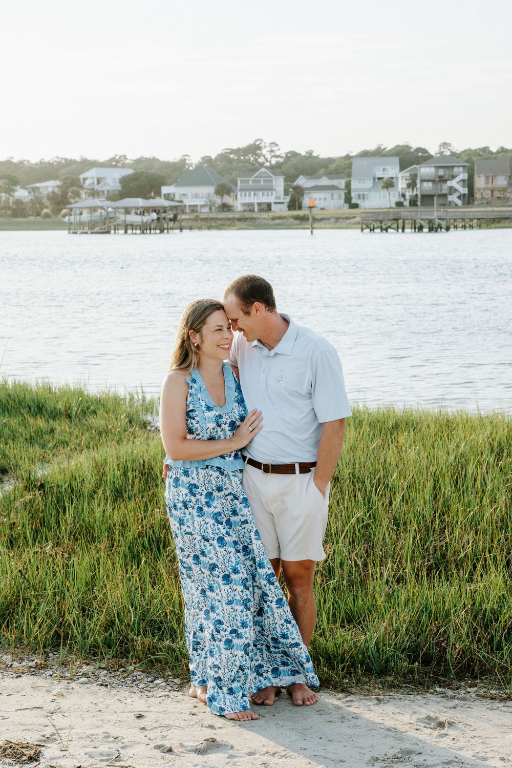 Couple embraces on sandy shore with marsh grass, water, and coastal homes in background.