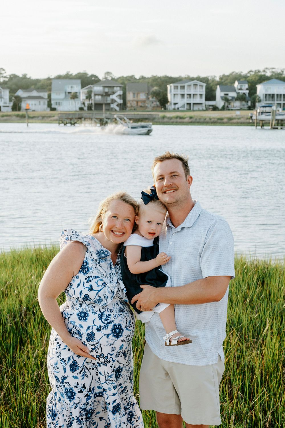 Happy family of three posing by waterfront, mother in floral dress, father holding baby girl in black outfit.