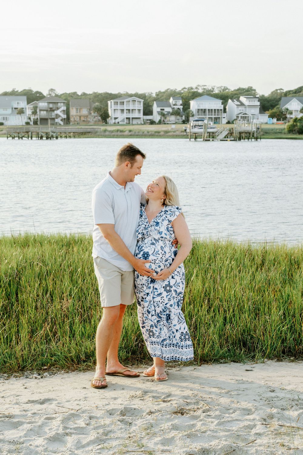 Couple standing on sandy beach by water, man holding pregnant woman's belly while smiling at each other near green marsh grass.