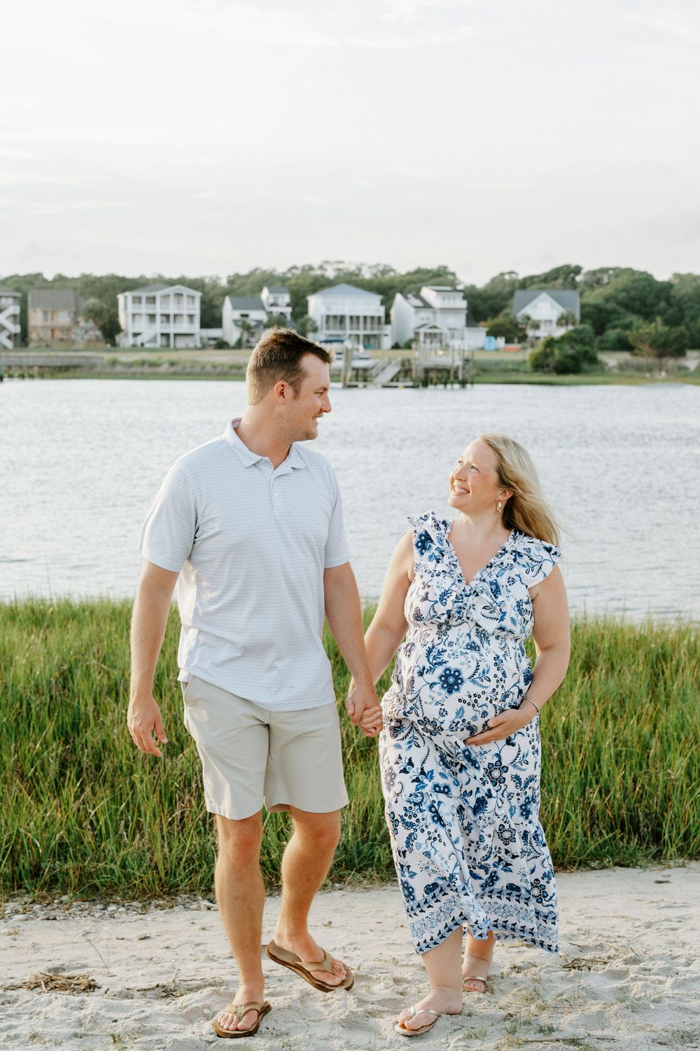 Pregnant woman in blue floral dress holds hands with man in white polo by waterfront beach.