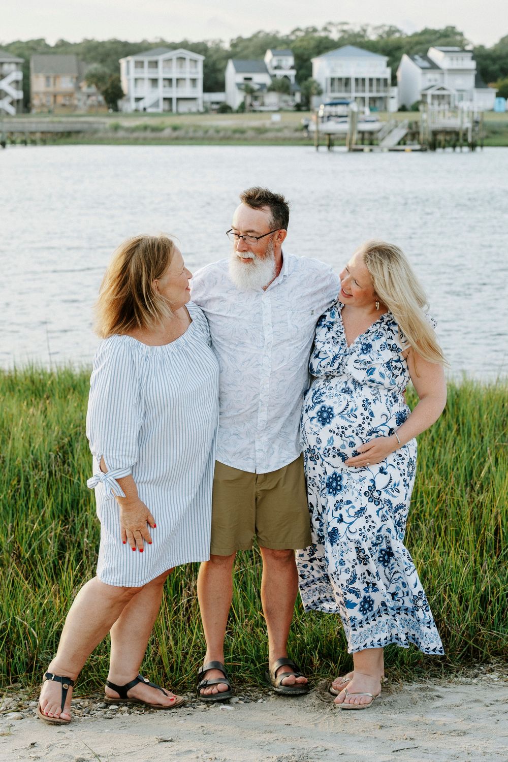 Three adults smiling together on a sandy beach with marsh grass and waterfront homes in the background.