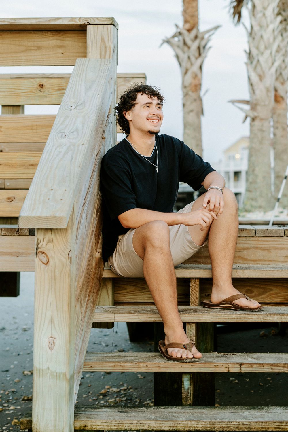 Smiling young man with curly hair sitting on wooden beach steps wearing black shirt and khaki shorts on cloudy day.