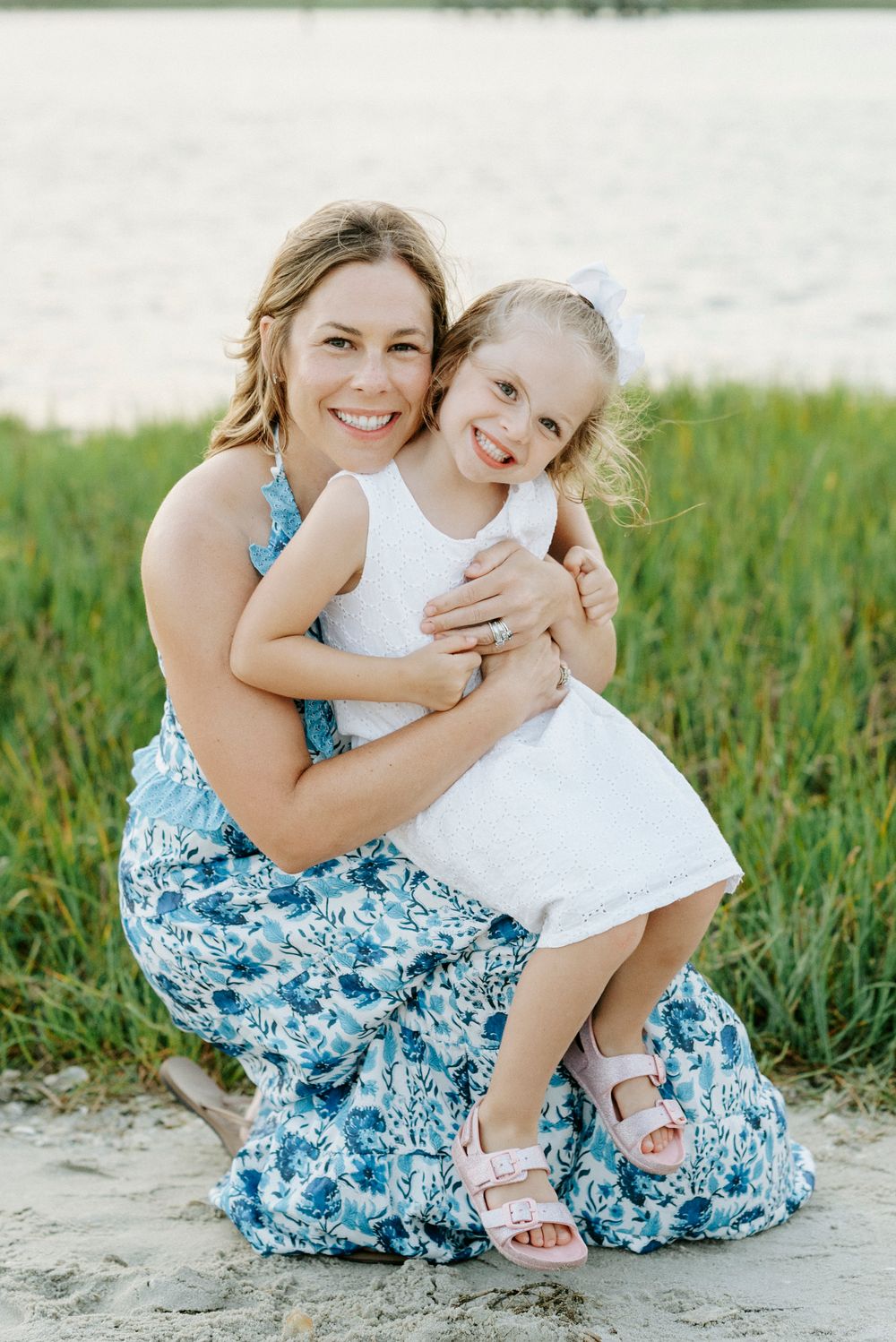 Smiling mother in blue floral dress hugs young blonde girl in white dress on sandy beach.