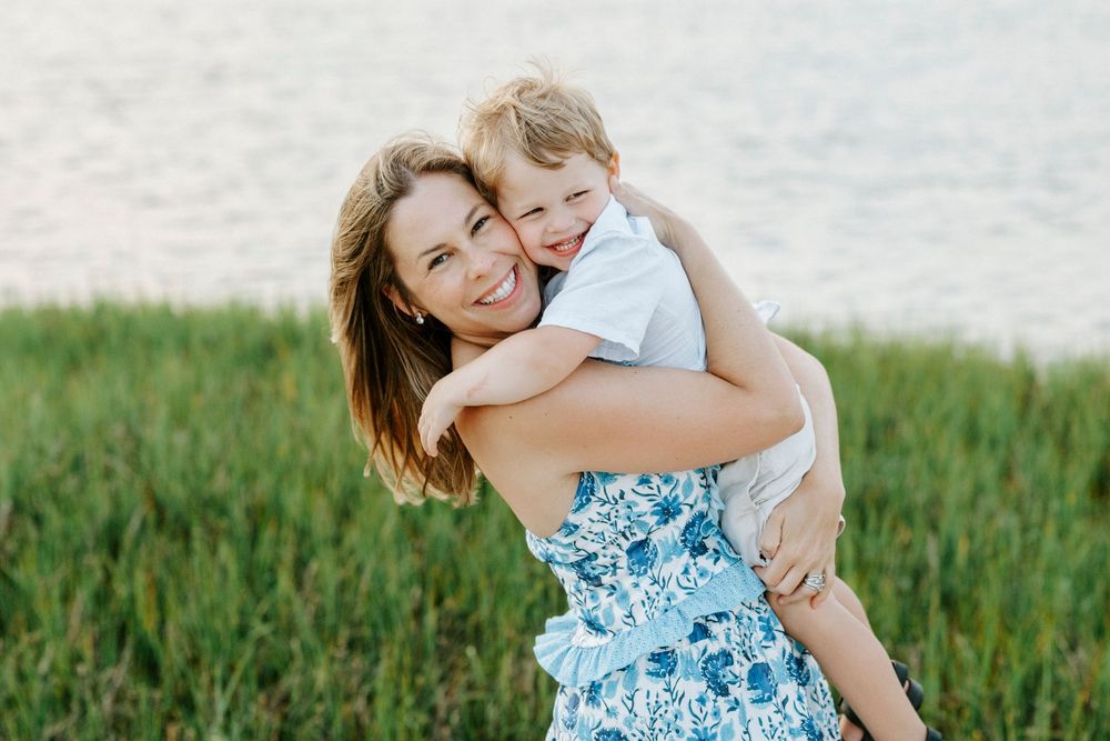 Happy brunette mother in blue floral dress hugs toddler boy near grassy waterfront.