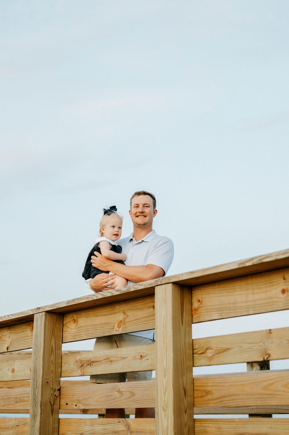 A smiling man holds a baby girl in a black outfit on a wooden boardwalk railing against a cloudy sky.