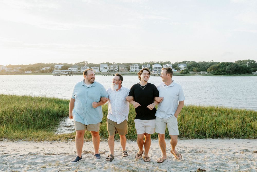Four men laughing together on sandy beach with marsh grass and waterfront homes in background.
