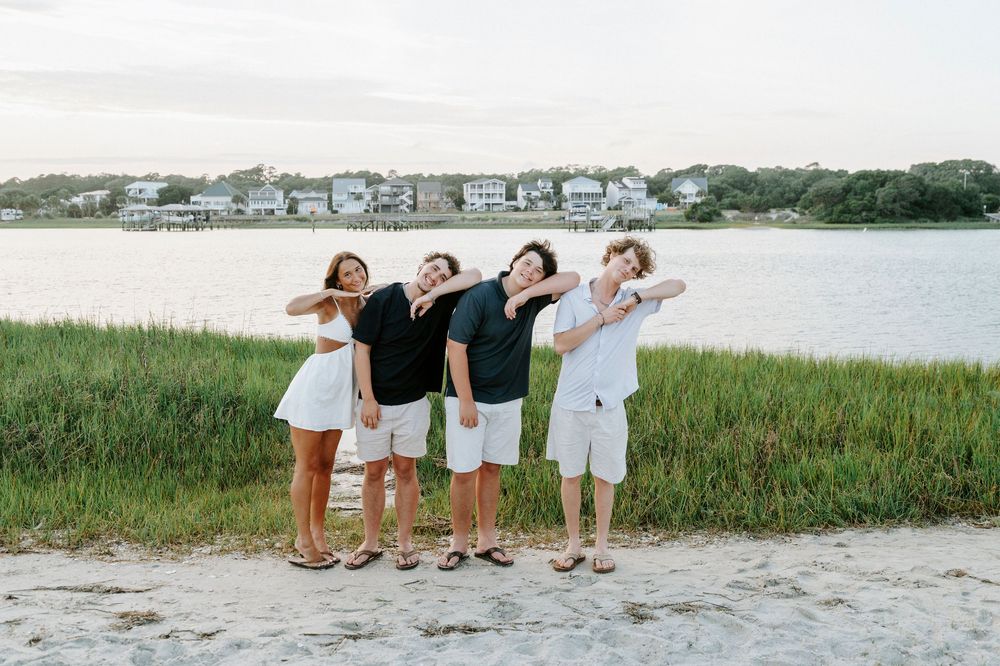 Four teens pose playfully on sandy beach with waterfront homes in background at sunset.