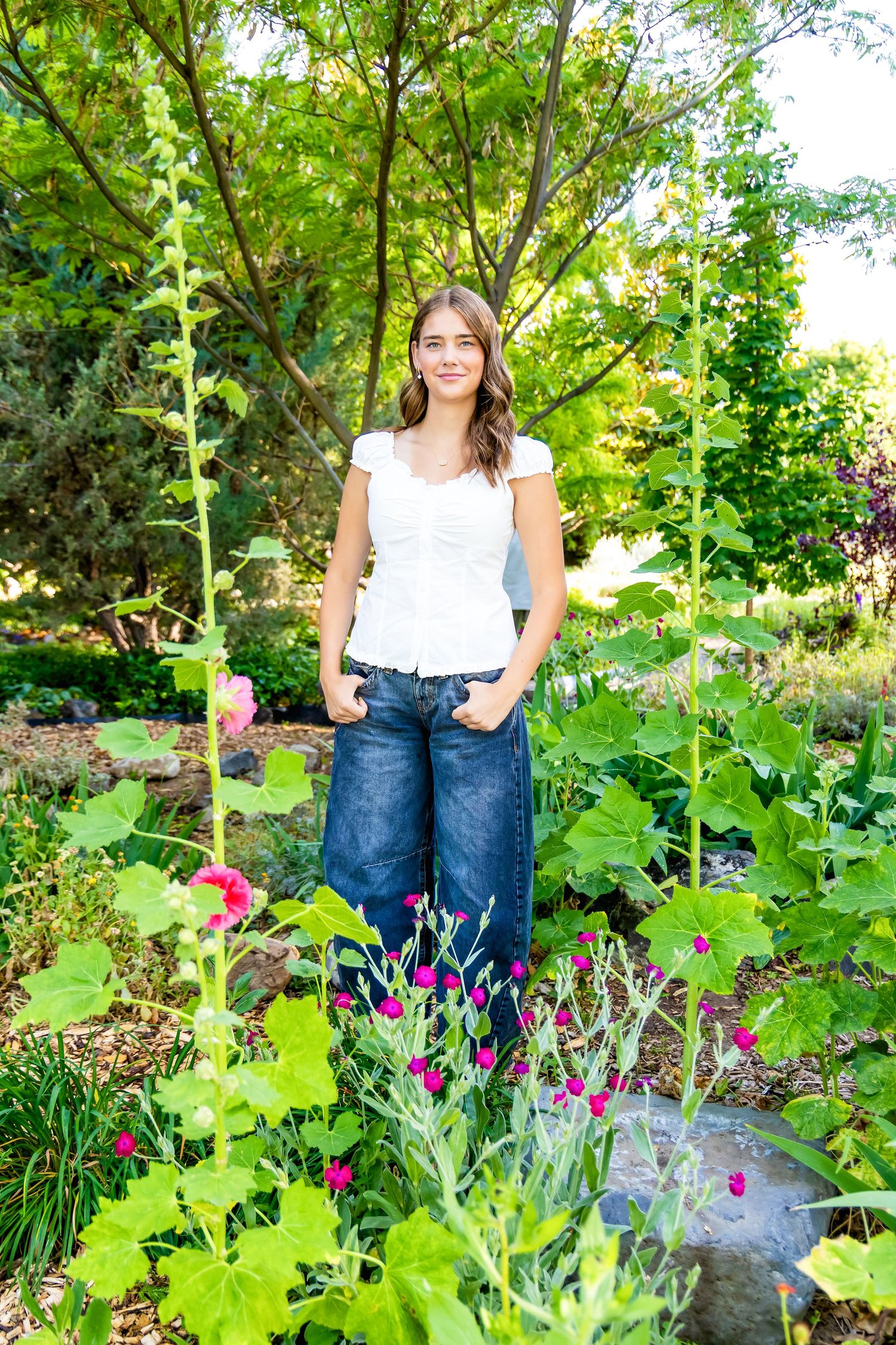 Young woman in white top and jeans smiling in a lush garden surrounded by colorful flowers and green foliage.