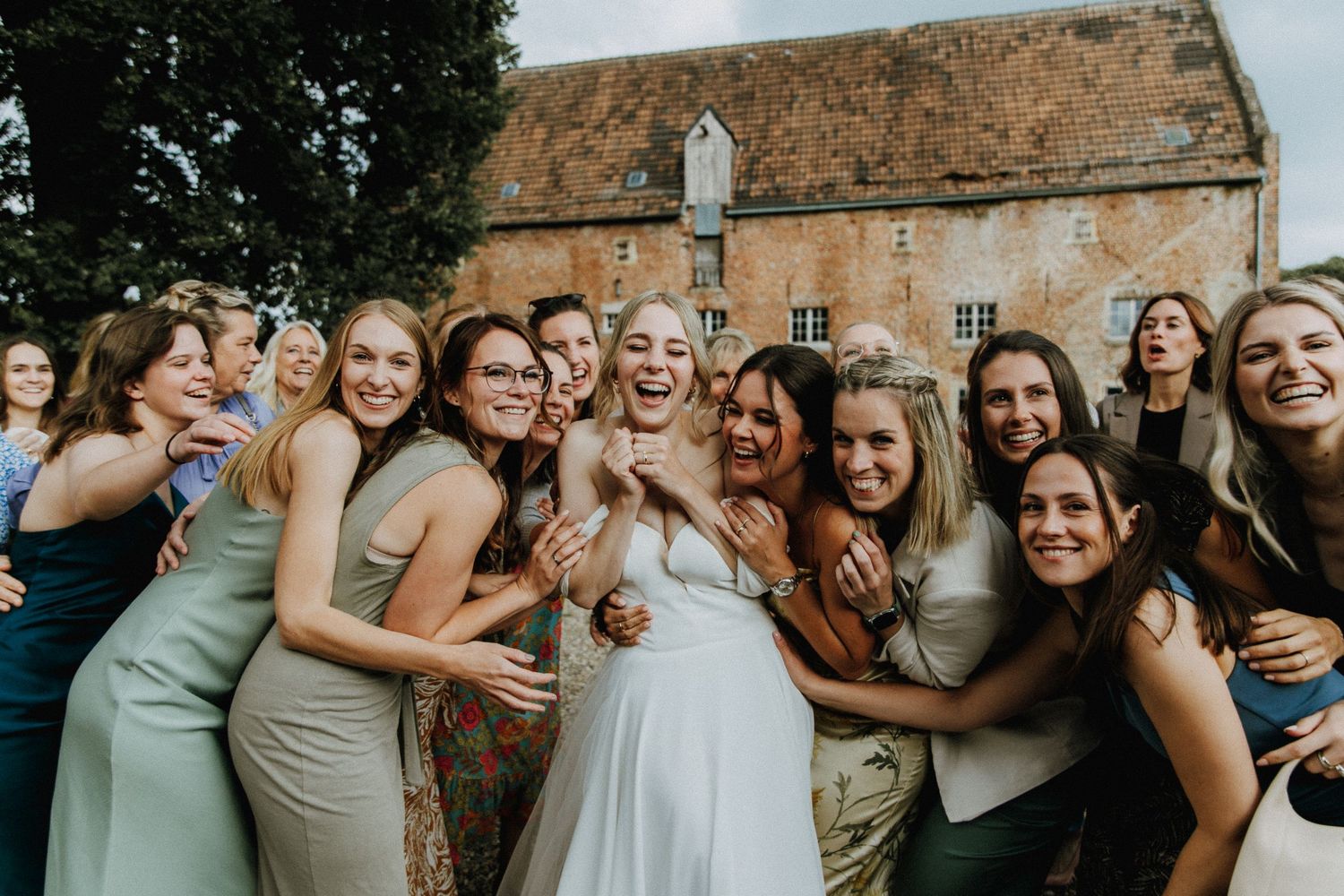 Bride in white gown surrounded by smiling bridesmaids in sage green dresses, celebrating outdoors near a historic stone building.