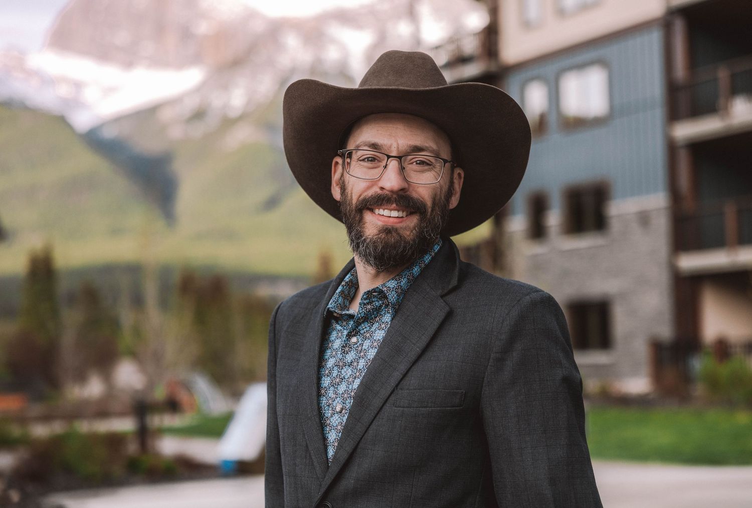 Smiling bearded man in cowboy hat and blazer poses outdoors with mountain backdrop.
