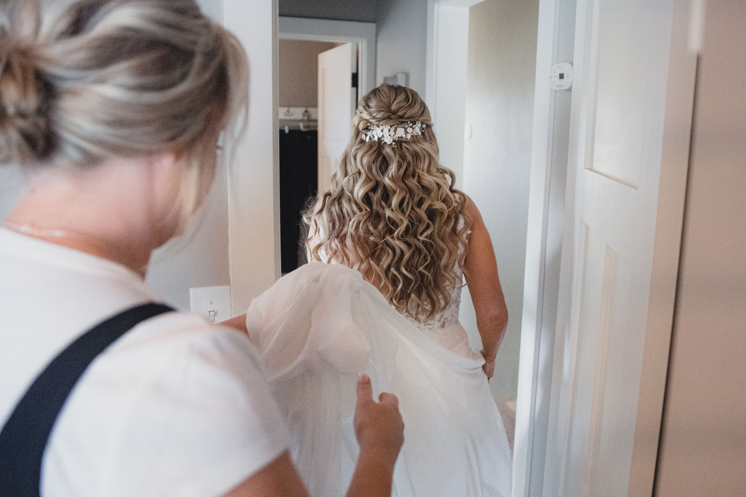 Bride in white wedding gown with curly hair and tiara being helped by bridesmaid in bridal suite.
