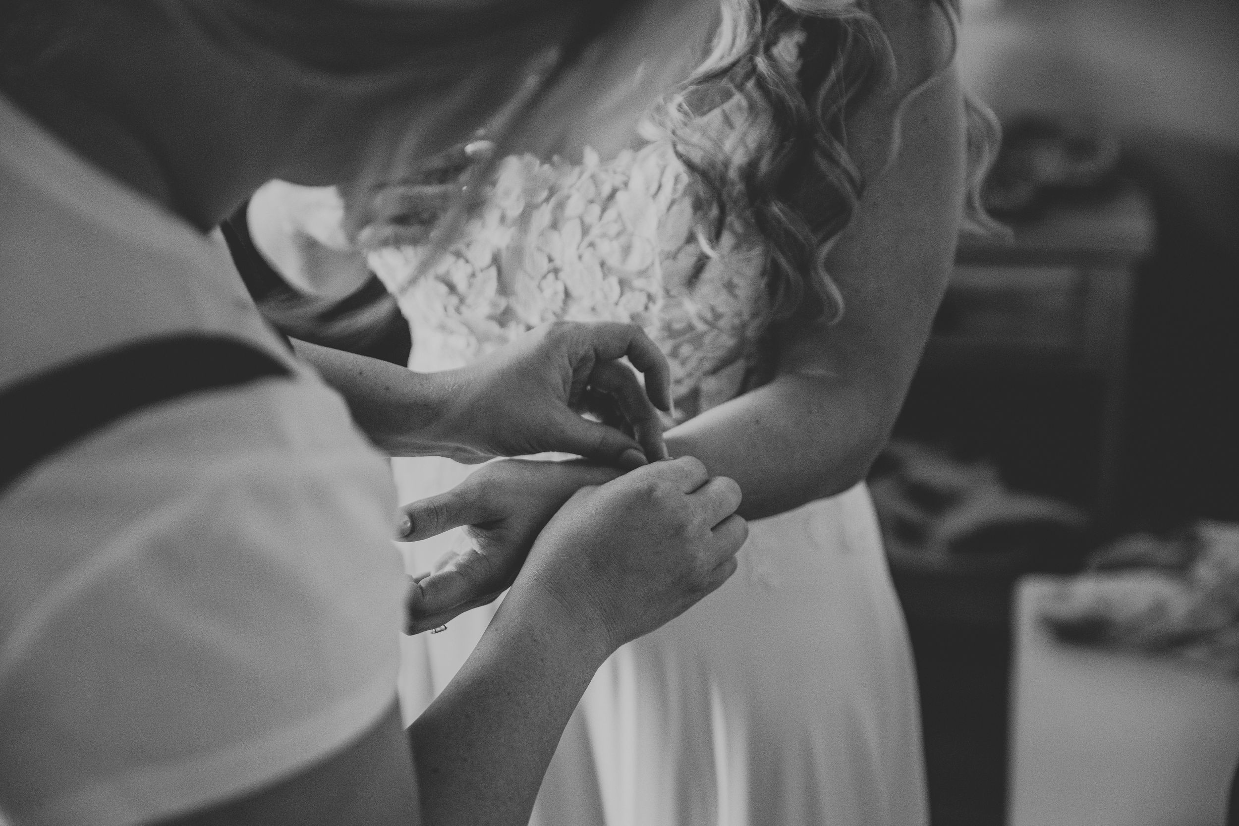 Black and white photo of a bride getting her wedding dress fastened by an assistant during bridal preparations.