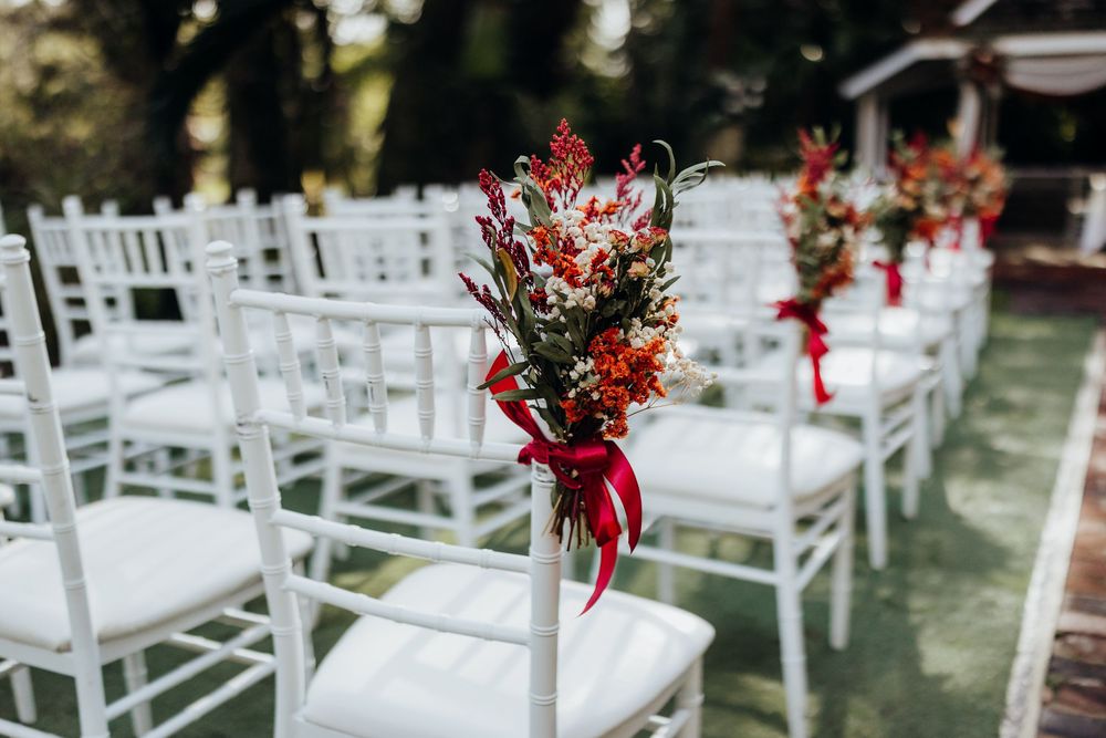 White chiavari chairs with red floral arrangements at outdoor wedding ceremony.