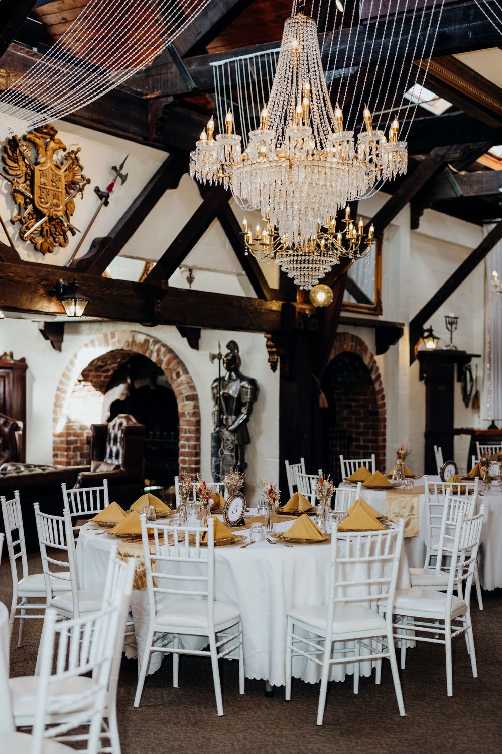Elegant wedding reception hall with crystal chandelier, white Chiavari chairs, round tables with gold napkins, and medieval armor decor.