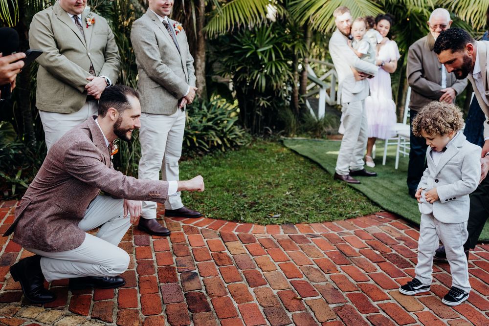 Groom crouches to greet young child in suit at outdoor wedding ceremony on brick path.