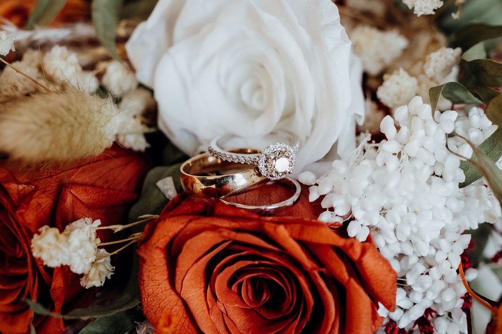 Gold wedding bands and diamond engagement ring resting on a bouquet of red and white roses with baby's breath.