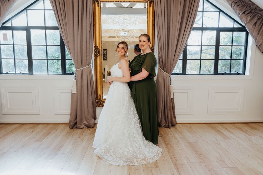 Bride in white lace gown posing with bridesmaid in olive green dress in bright wedding venue.