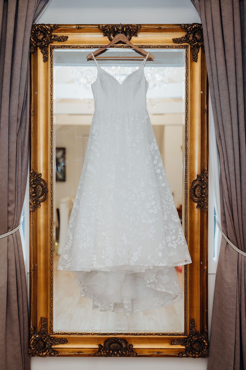 White lace A-line wedding dress hanging on a wooden hanger in an ornate gold mirror frame.