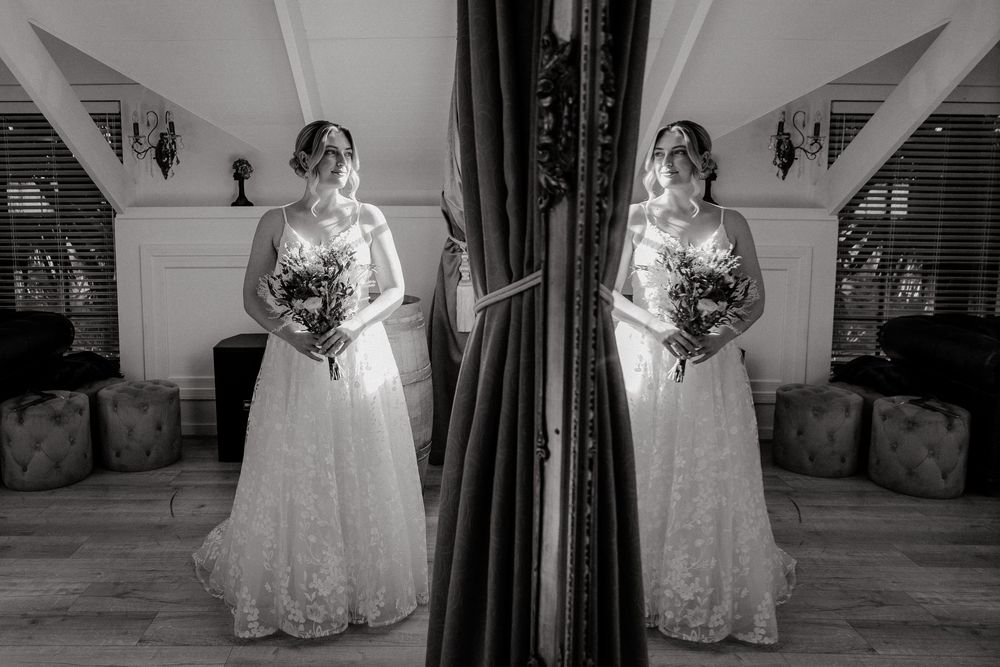 Black and white portrait of a bride in lace gown holding bouquet, reflected in a mirror before her wedding ceremony.