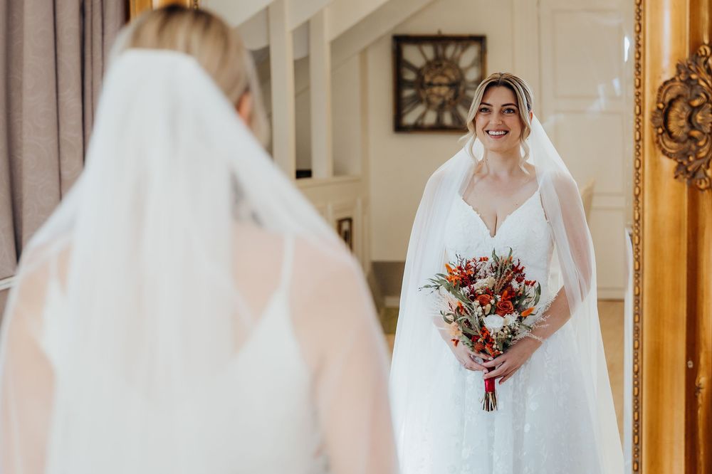 Smiling bride in white gown and veil holds colorful bouquet while viewing her reflection in an ornate mirror.