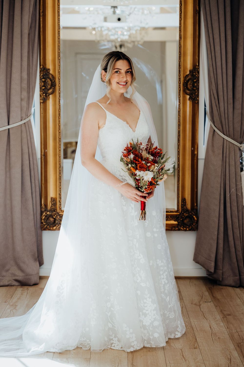 Bride in white lace A-line gown with veil holds red bouquet before ornate gold mirror.