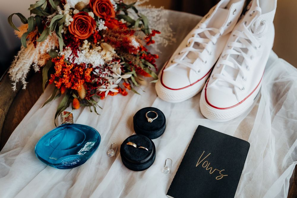 Wedding flat lay featuring red bouquet, white Converse sneakers, gold rings, blue dish, and vows booklet.
