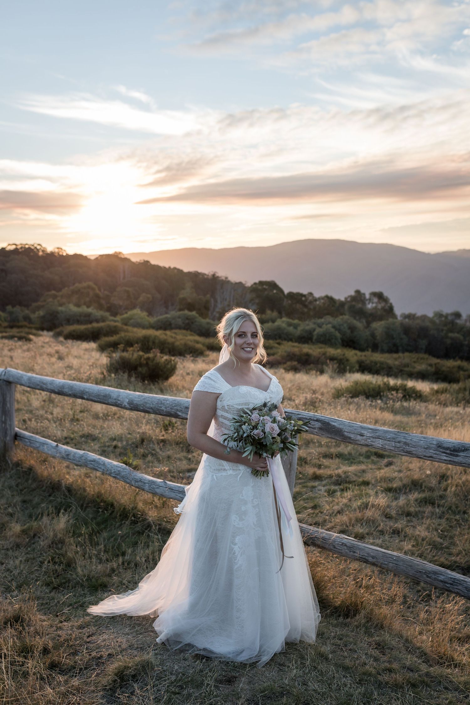Bride in white lace gown holds bouquet at sunset with mountain backdrop and rustic fence.