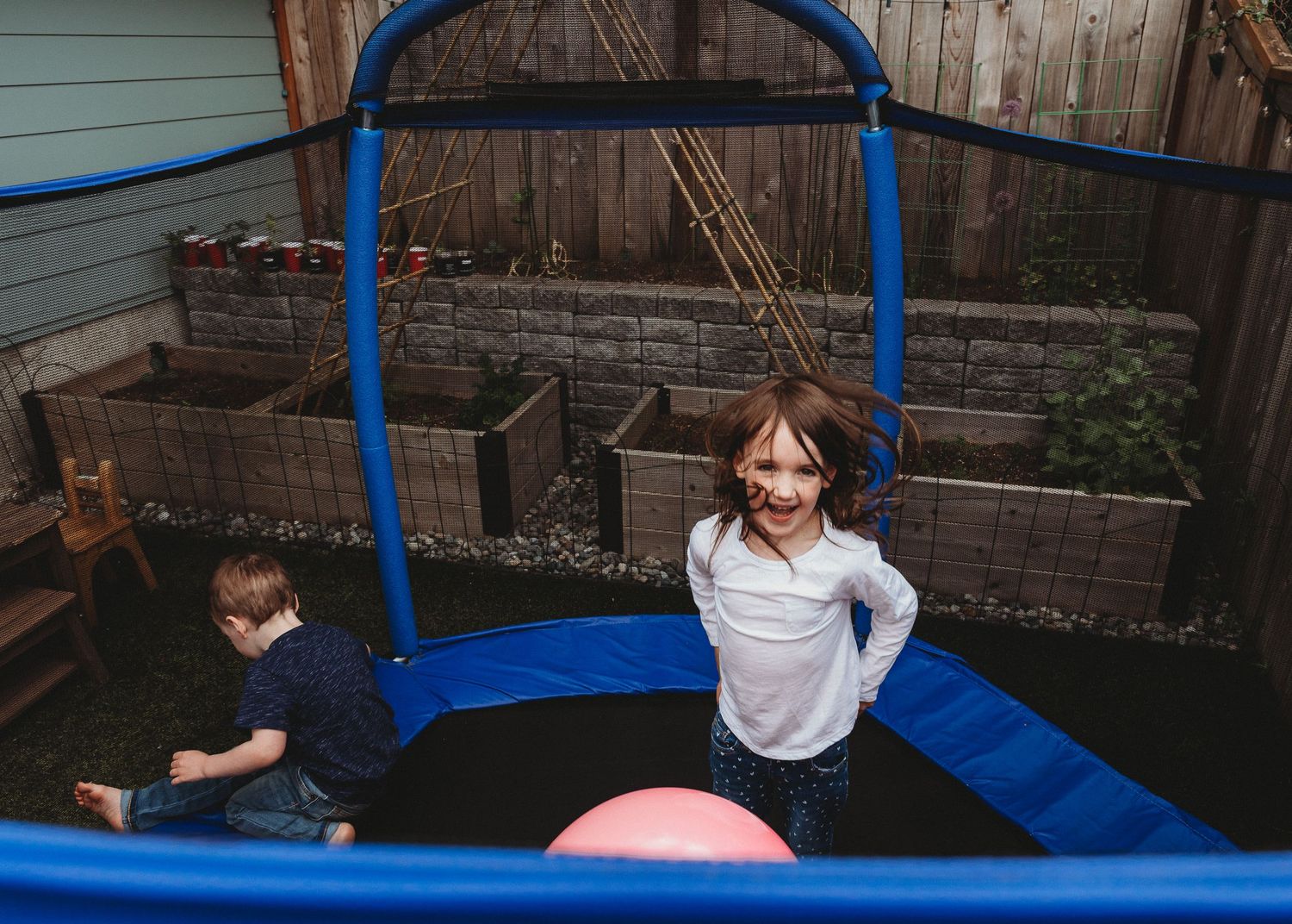 Two children playing on a blue trampoline in a backyard, girl smiling at camera, boy climbing up beside her.