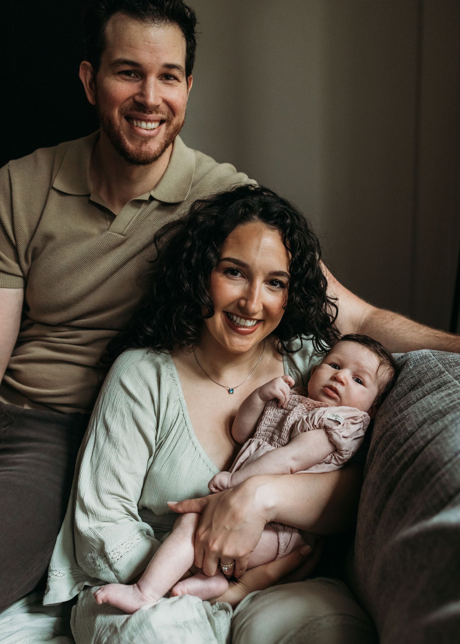 Smiling couple sitting on a couch holding their newborn baby, captured in a warm indoor family portrait.