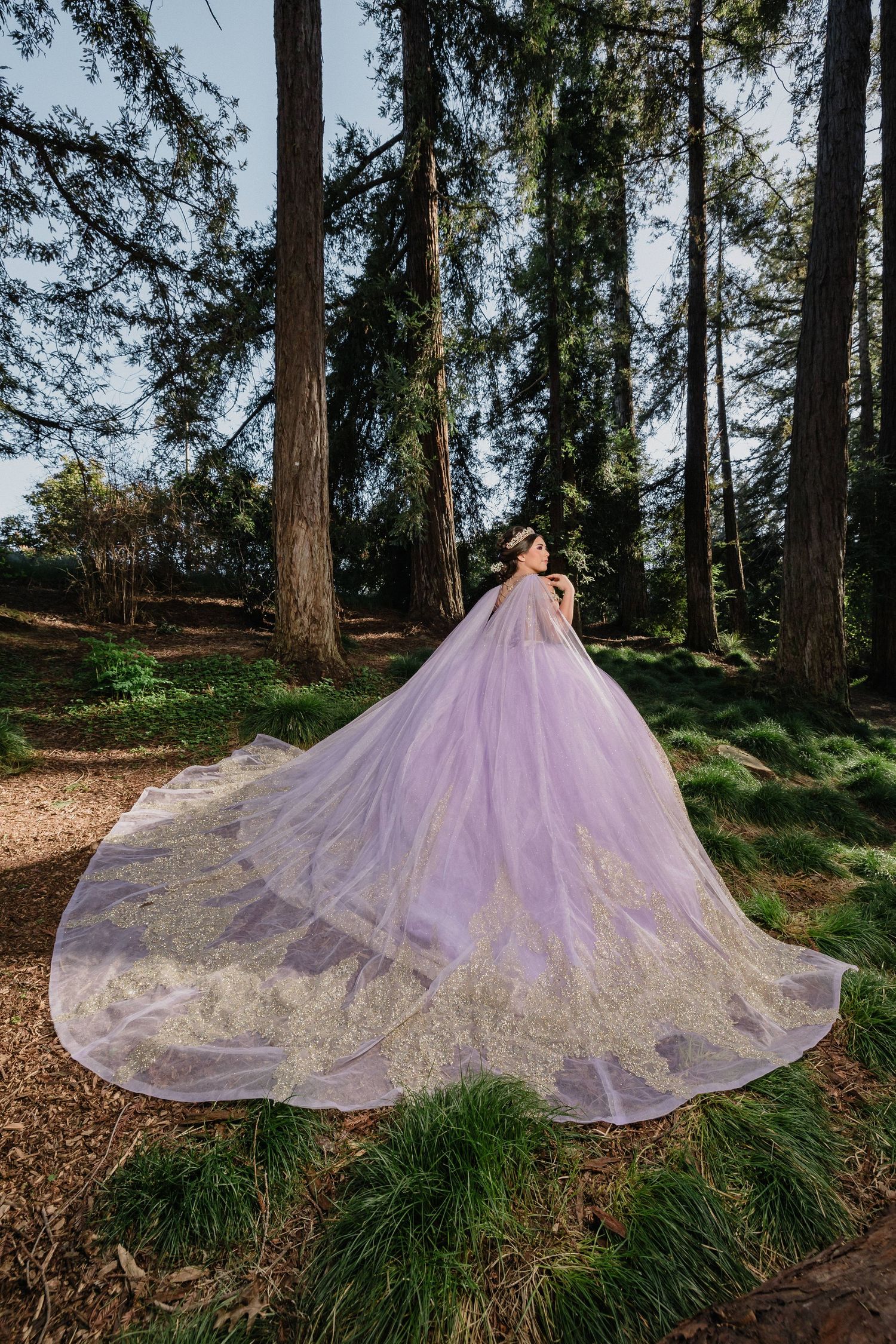 Woman in blue ball gown poses by a pond in front of a white castle-like mansion on a sunny day.